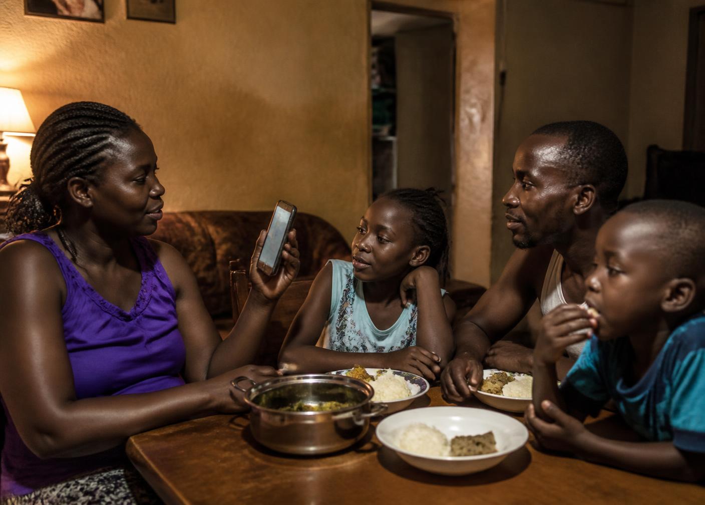 A family in a Kampala home checking a bill on a phone over an evening meal